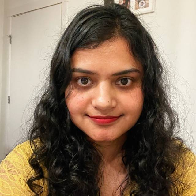 A person with long, curly dark hair smiles at the camera. They are wearing a yellow lace top, and the background includes a white door and a small picture frame on the wall.