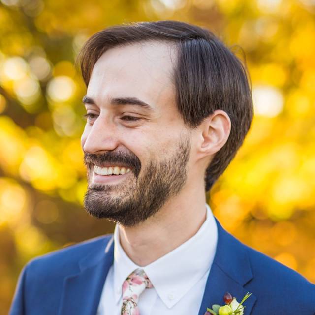 A man in a blue suit with a floral tie and a boutonniere smiles while looking to the side. The background features bright, blurred autumn foliage in shades of yellow and gold.