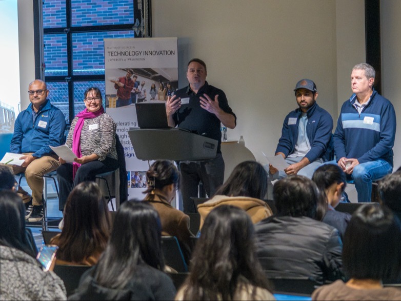 A panel of five people, four men and one woman, sit in front of an audience. The person at the podium is speaking. A banner behind them reads Technology Innovation. The audience is facing the panel in a lecture-style setting.
