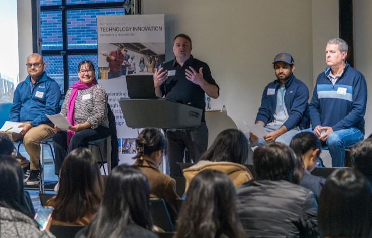 A panel of five people, four men and one woman, sit in front of an audience. The person at the podium is speaking. A banner behind them reads Technology Innovation. The audience is facing the panel in a lecture-style setting.