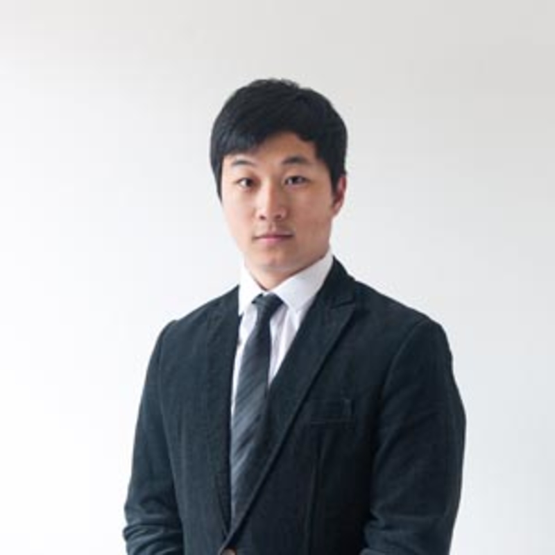 A man in a black suit and tie stands against a plain white background, looking directly at the camera. He has short black hair and a neutral expression.
