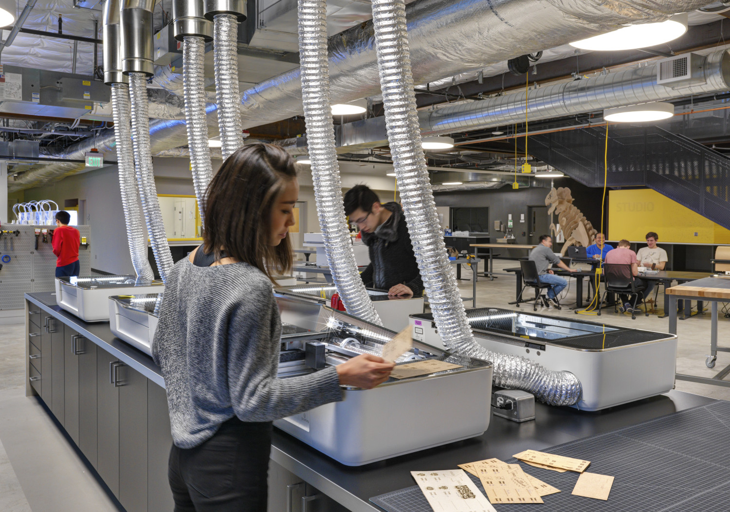 A woman works at a laser cutting station in a tech workshop. Several vent hoses are connected to the machines. In the background, people interact at tables. The space has an industrial look with exposed ducts and high ceilings.