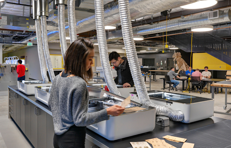 A woman works at a laser cutting station in a tech workshop. Several vent hoses are connected to the machines. In the background, people interact at tables. The space has an industrial look with exposed ducts and high ceilings.
