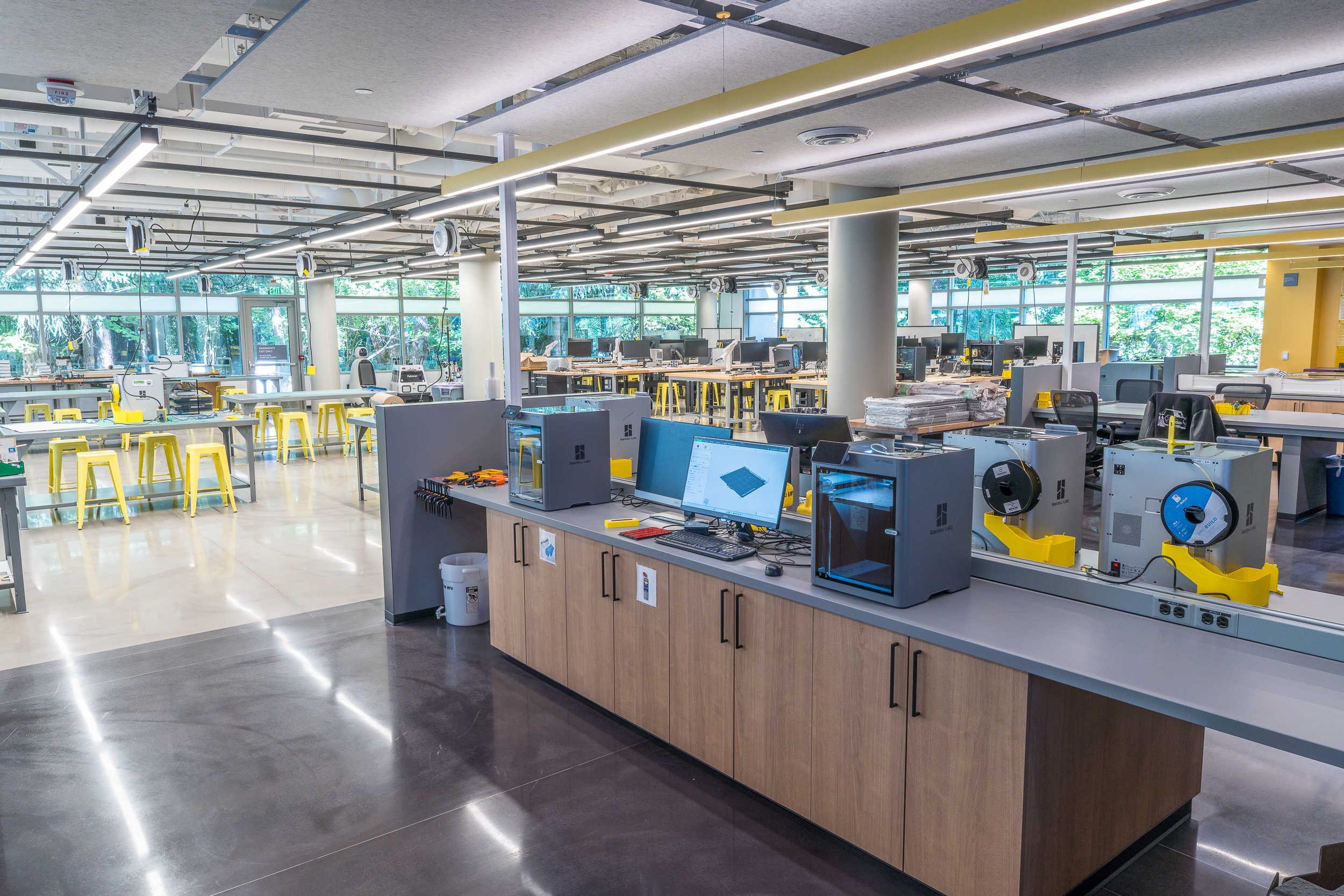 A woman works at a laser cutting station in a tech workshop. Several vent hoses are connected to the machines. In the background, people interact at tables. The space has an industrial look with exposed ducts and high ceilings.