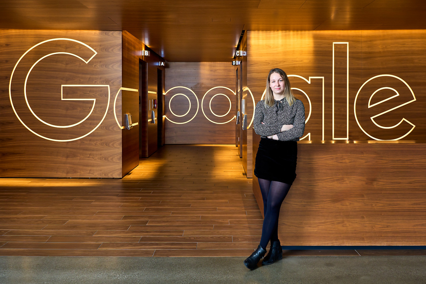 A person stands with arms crossed in front of a wooden wall with the word Google outlined in light. The corridor behind leads to multiple doors, and the space has a modern aesthetic.