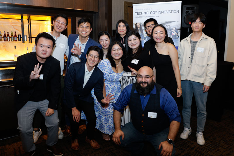 A group of people are posing and smiling together in a room with a bar in the background. They are dressed in semi-formal attire, and a poster about technology innovation is partially visible behind them.