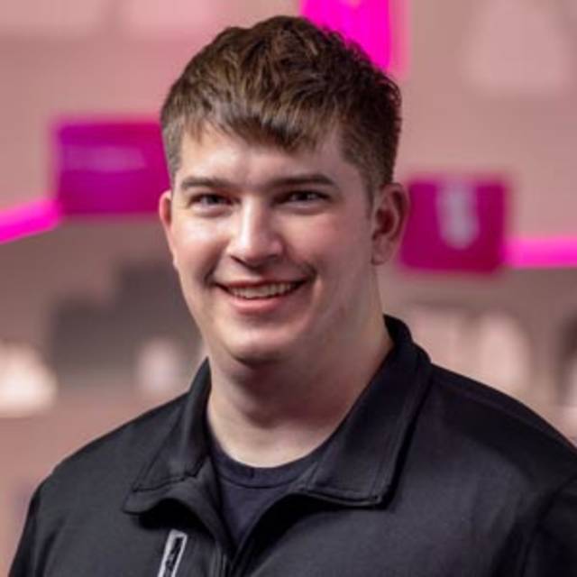 A person with short brown hair is smiling. They are wearing a black jacket over a dark shirt. The background is blurred with shades of pink and gray.