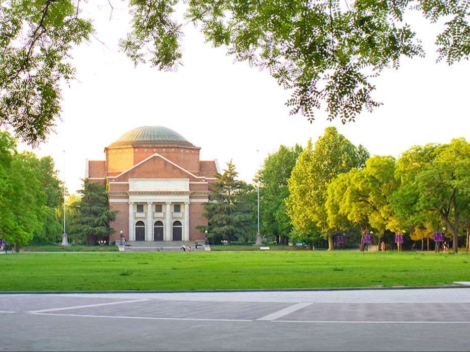 A historic red brick building with a large dome sits in a spacious grassy area surrounded by lush green trees. The building features tall columns at the entrance and is set against a clear sky. A paved walkway leads toward it.