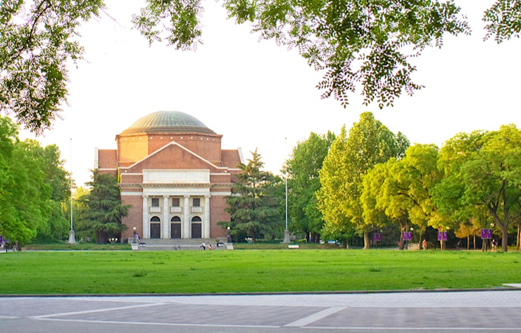 A historic red brick building with a large dome sits in a spacious grassy area surrounded by lush green trees. The building features tall columns at the entrance and is set against a clear sky. A paved walkway leads toward it.