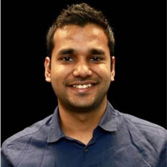 A person with short dark hair is smiling, wearing a dark button-up shirt, and looking at the camera against a black background.