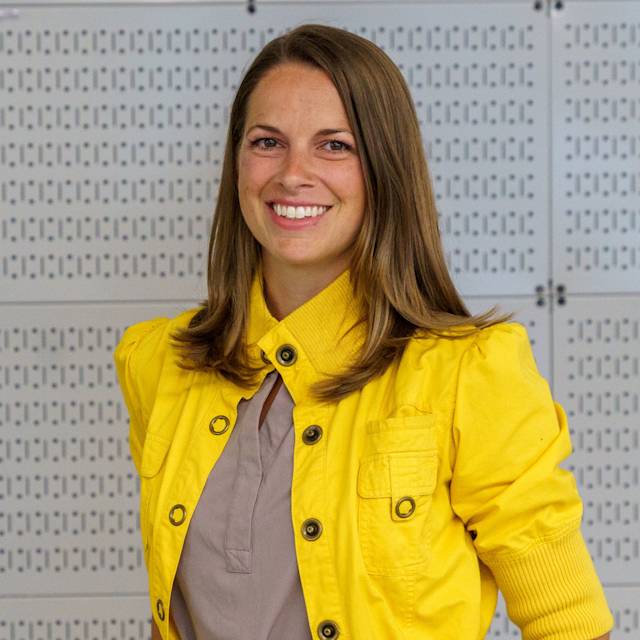 A person with long brown hair, wearing a bright yellow jacket over a beige top, smiles while standing against a patterned grey wall.