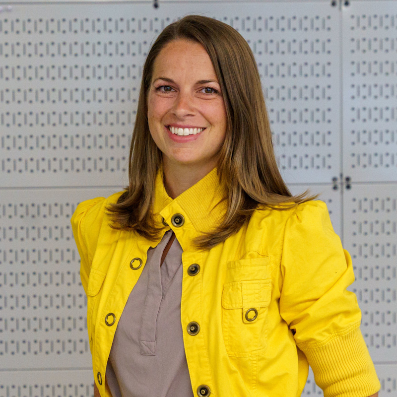A person with long brown hair, wearing a bright yellow jacket over a beige top, smiles while standing against a patterned grey wall.