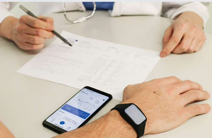 A person points at a document with a pen, which is on a table. Another person is wearing a smartwatch and has a smartphone in front of them displaying charts and data. Both are seated at the table.