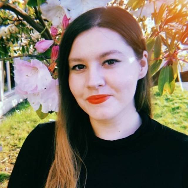 A person with long brown hair and red lipstick smiles in front of blooming pink flowers and green leaves. They are wearing a black top, and the background shows sunlight filtering through foliage.