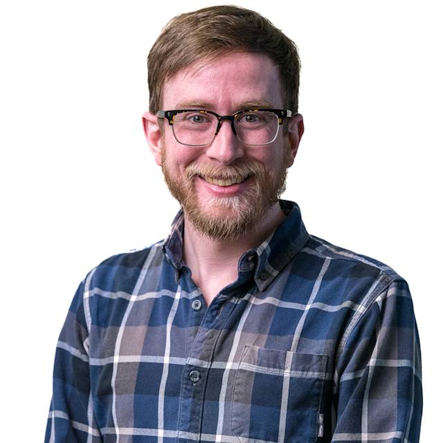A person with short brown hair and glasses smiling, wearing a black and white plaid shirt. They are standing with arms crossed in front of a patterned background.