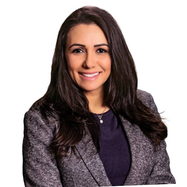 A person with long, dark hair is smiling, wearing a gray blazer over a black top, and a necklace. The background is plain white.