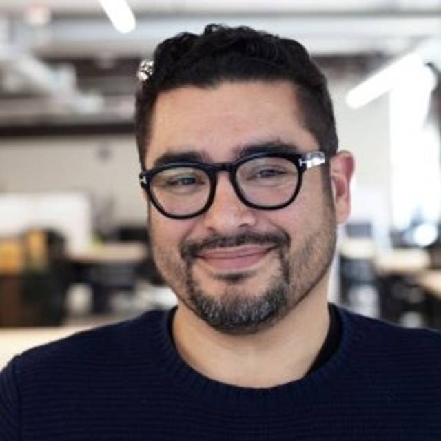 Person smiling indoors, wearing glasses and a dark sweater. The background features an office setting with blurred lights and furniture.