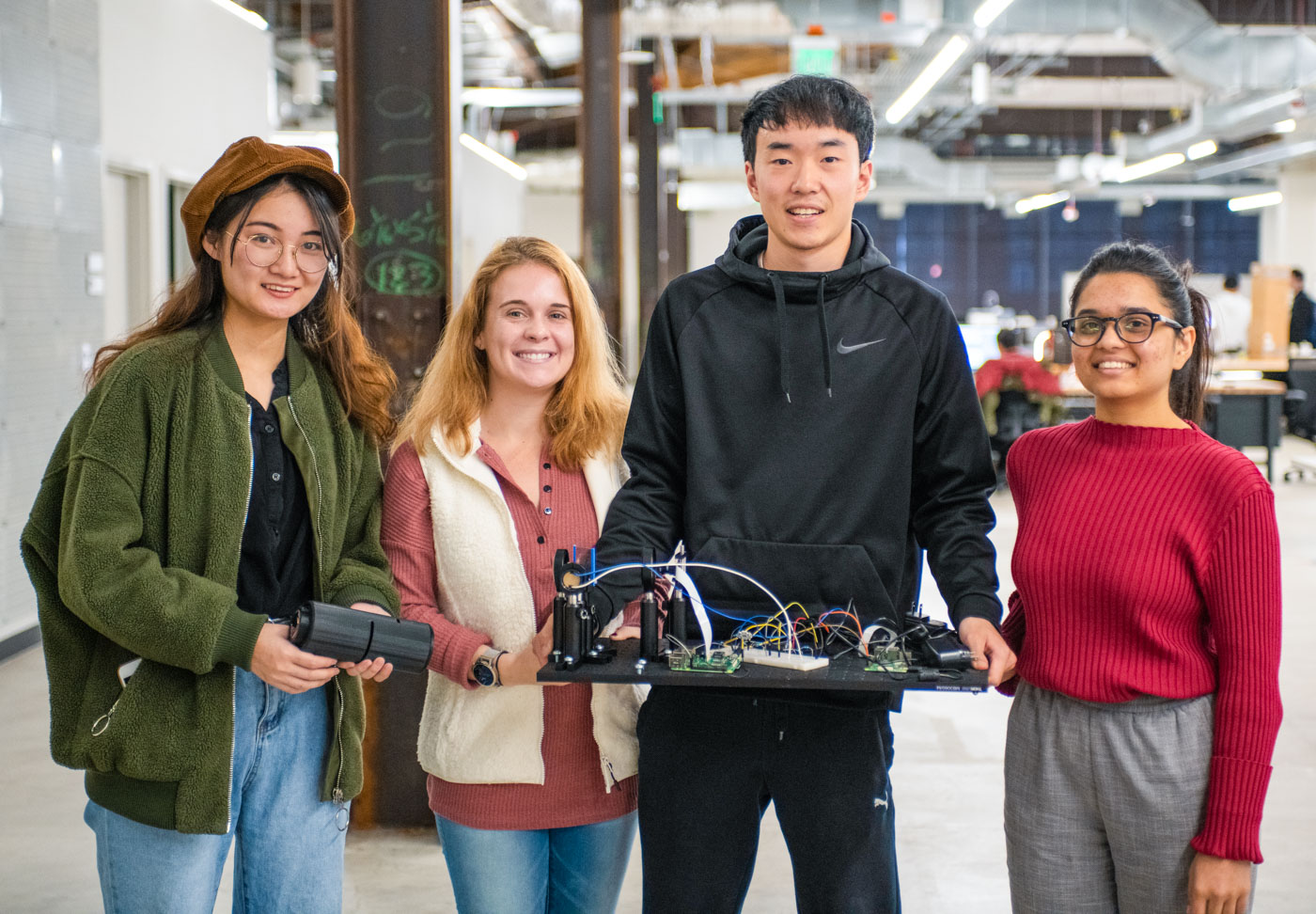 Four people stand inside a modern office space, smiling at the camera. They hold a circuit board project with various wires connected. The office features exposed ductwork and industrial design elements.