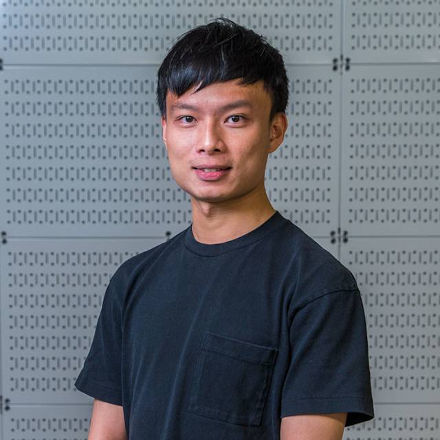 A person with short black hair wearing a black t-shirt stands in front of a wall with a repetitive pattern.