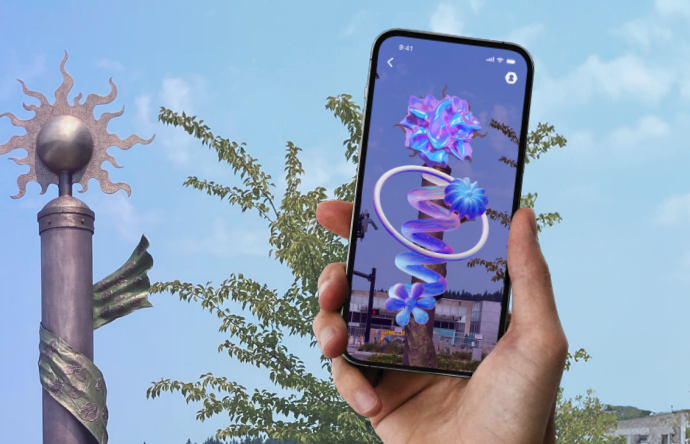 A hand holds a smartphone displaying an augmented reality sculpture with swirling flowers and rings. The background shows a metal pole with a sun sculpture on top, and leafy branches against a blue sky.