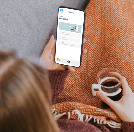 A person with long hair is holding a smartphone displaying a screen titled Therapy: Needles with checkboxes. They are sitting covered with a herringbone-patterned blanket, holding a mug of coffee in the other hand.