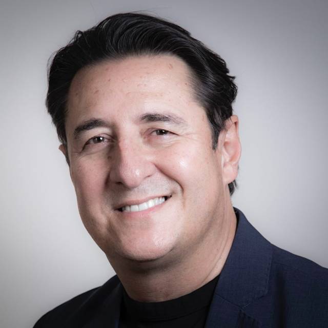 A person with dark hair, wearing a dark suit jacket and black shirt, smiles at the camera against a plain background.
