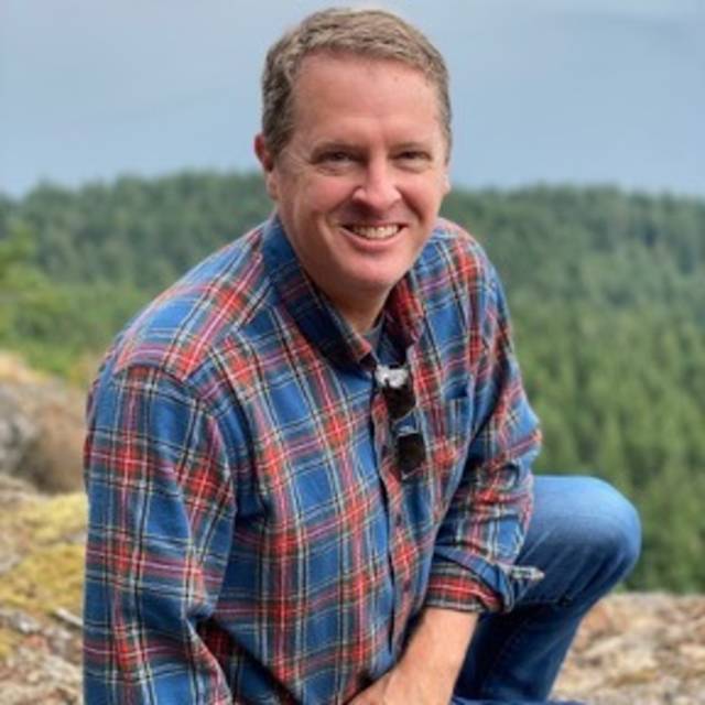 A person wearing a blue and red plaid shirt kneeling on a rocky surface with a forested landscape in the background. They are smiling at the camera.
