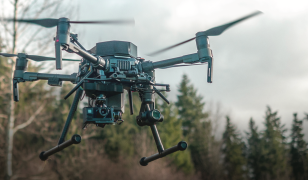 A drone with a mounted camera flies in an outdoor setting. The drone is black, featuring four propellers, and appears to be navigating through a forested area with trees visible in the background against a cloudy sky.
