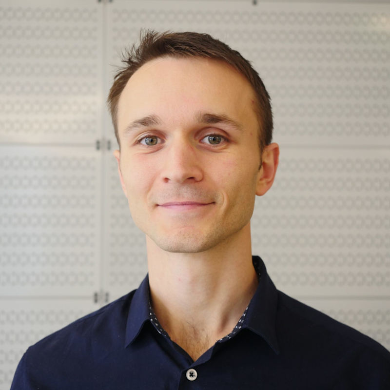 A person with short brown hair wearing a dark navy blue shirt stands against a light-colored, patterned background, looking at the camera and smiling gently.