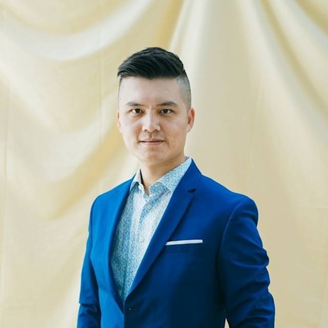A person wearing a blue suit and a white patterned shirt stands against a light beige draped background, looking directly at the camera with a neutral expression.