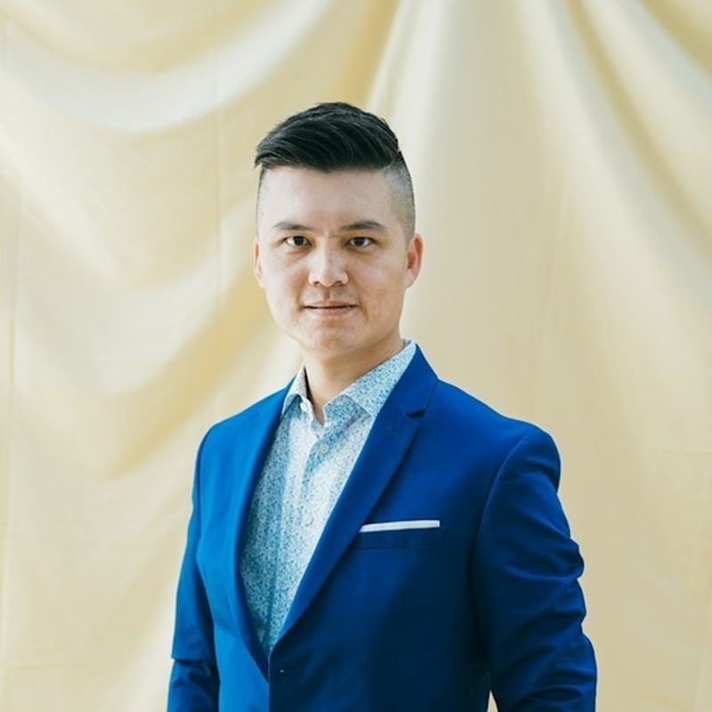 A person wearing a blue suit and a white patterned shirt stands against a light beige draped background, looking directly at the camera with a neutral expression.