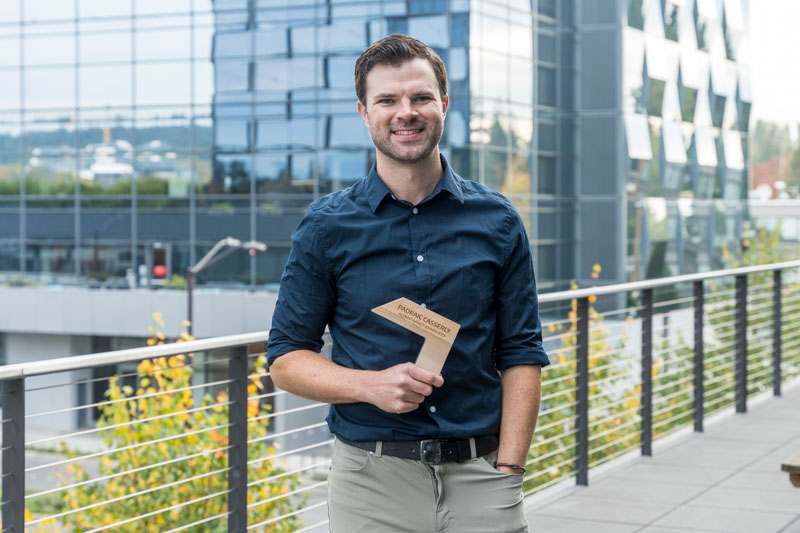 A man in a dark shirt and light pants stands on an outdoor balcony, smiling and holding a triangular wooden award. Behind him is a modern glass building with geometric patterns.