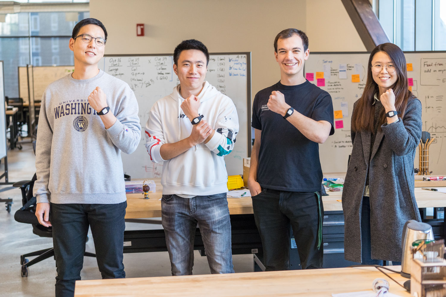 Four people standing in an office, smiling and showing off smartwatches on their wrists. They are in front of a whiteboard filled with notes and diagrams. One person wears a University of Washington sweatshirt. Casual attire.