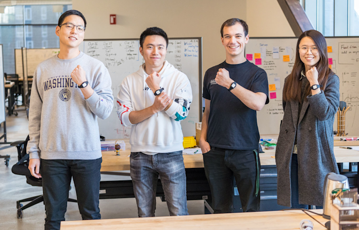 Four people standing in an office, smiling and showing off smartwatches on their wrists. They are in front of a whiteboard filled with notes and diagrams. One person wears a University of Washington sweatshirt. Casual attire.