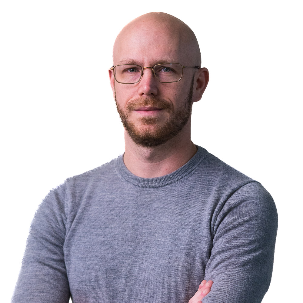A person with a bald head and beard wearing glasses and a dark polo shirt stands with arms crossed. The background consists of a perforated metal surface with a grid pattern.