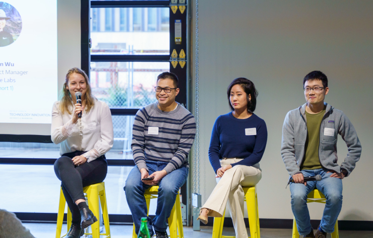 Four people sit on yellow stools in a panel discussion. One woman holds a microphone while smiling. A presentation slide is partially visible in the background. Casual attire and a relaxed atmosphere.