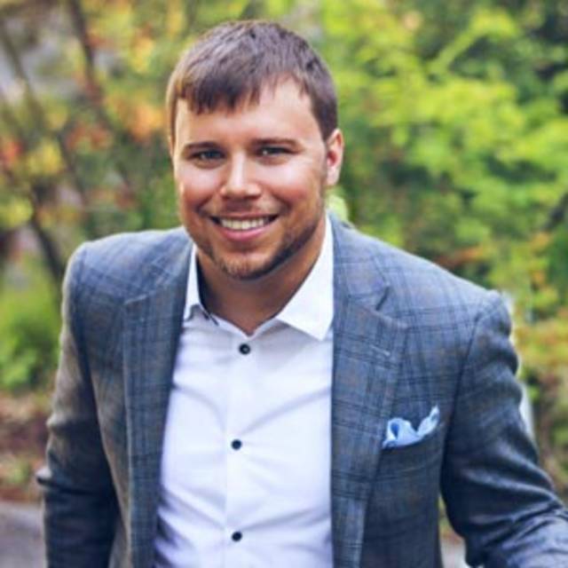 A man with short brown hair and a beard, wearing a gray plaid blazer, a white shirt, and a light blue pocket square, smiles at the camera. He is standing outdoors with green foliage in the background.