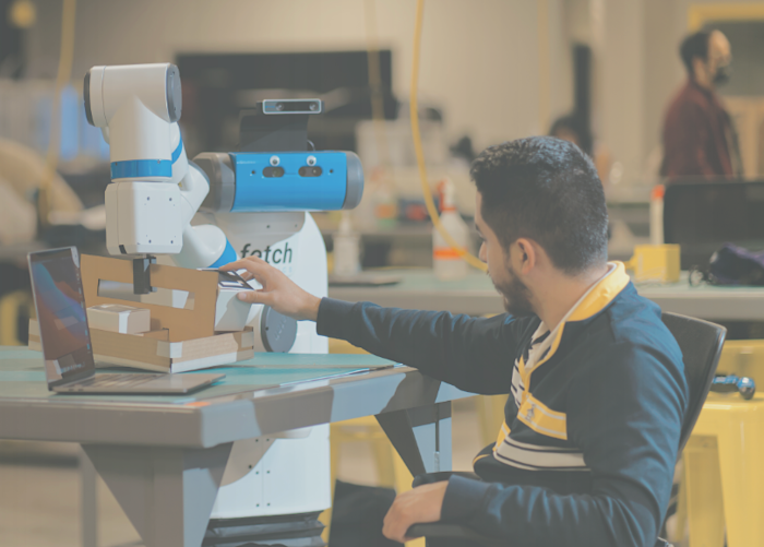 A person interacts with a robot arm, placing or adjusting an object on a table. The arm is labeled fetch. A laptop is open nearby, and another individual is blurred in the background. The setting appears to be a workshop or lab.