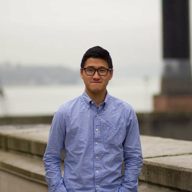 A person wearing glasses and a light blue button-up shirt stands outdoors with hands in pockets. The background features a blurred view of a body of water and a tall chimney on a cloudy day.