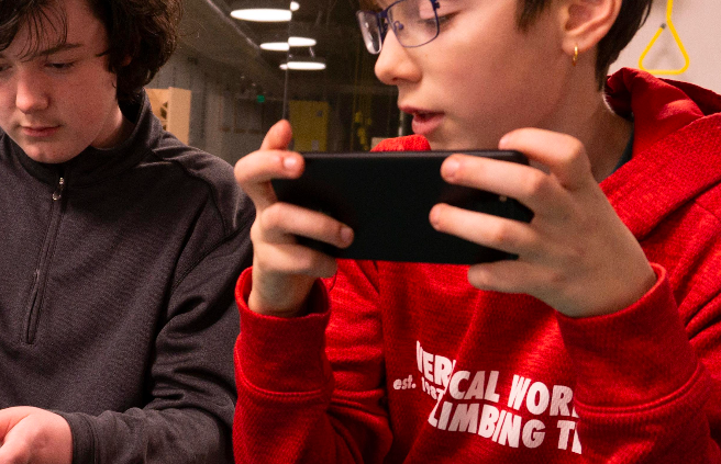 Two young people are indoors, focused on handheld devices. One is in a gray hoodie, looking down, and the other in a red hoodie, with glasses and an earring, holding a game controller. The background is softly lit with blurred details.