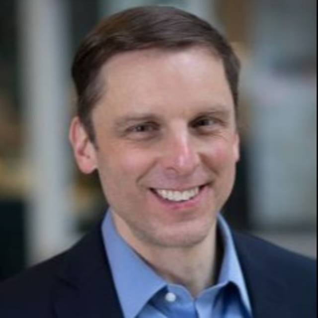 A person in a dark blazer and blue shirt smiles at the camera. The background is softly blurred, suggesting an indoor setting.