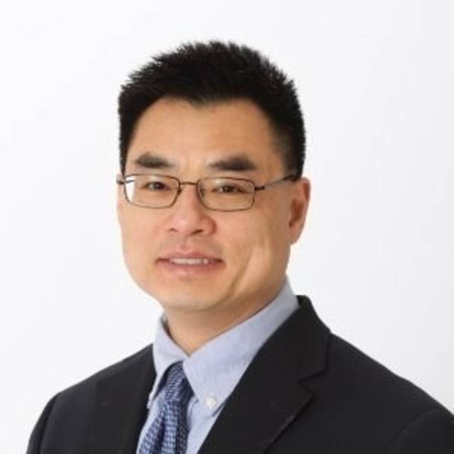 A person with short black hair and glasses wears a suit jacket and a blue button-up shirt with a tie, posing against a plain white background. They are smiling slightly.