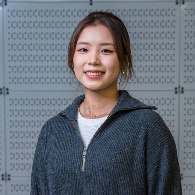A person with long hair smiles at the camera, wearing a gray zip-up sweater over a white shirt. The background features a wall with a geometric pattern.