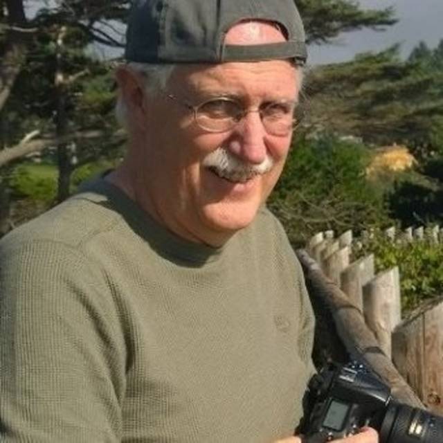 Older man in a gray cap and glasses, holding a camera. Hes outdoors with trees and a wooden fence in the background, smiling.