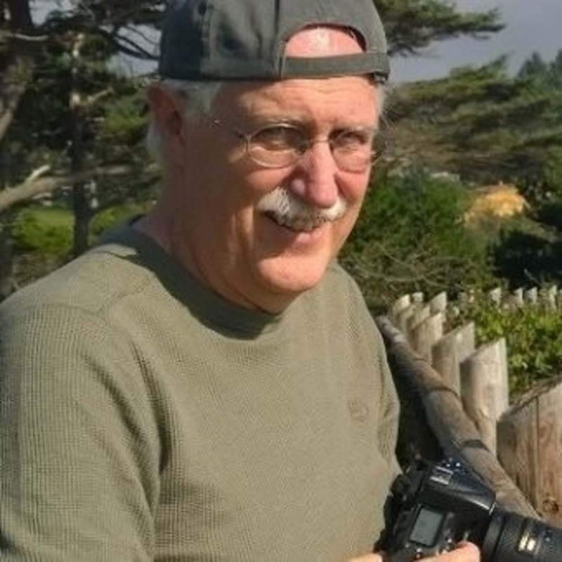 Older man in a gray cap and glasses, holding a camera. Hes outdoors with trees and a wooden fence in the background, smiling.