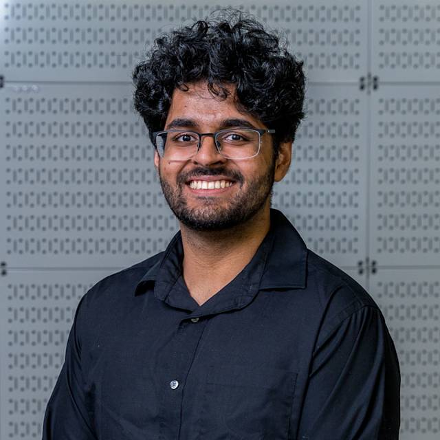 A person with curly hair, wearing glasses and a black shirt, is smiling at the camera. The background features a pattern of lines and squares.