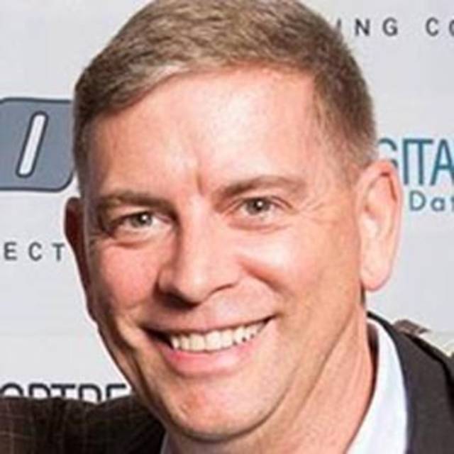 Smiling man with short hair wearing a suit jacket, photographed in front of a wall with text and logos.
