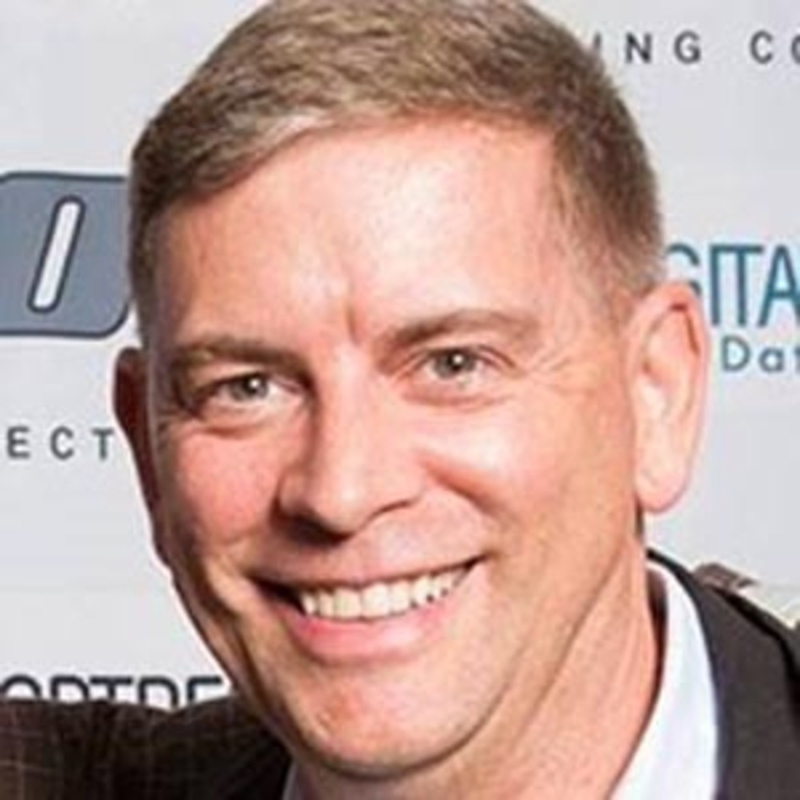 Smiling man with short hair wearing a suit jacket, photographed in front of a wall with text and logos.