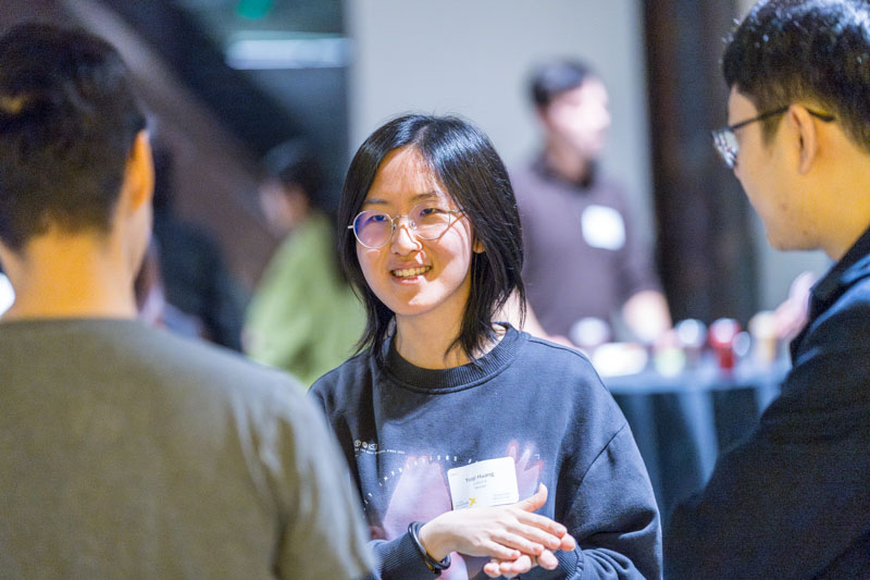 A person wearing glasses and a dark sweater is smiling and talking with two others at an indoor event. There are blurred people and tables in the background, indicating a lively gathering.