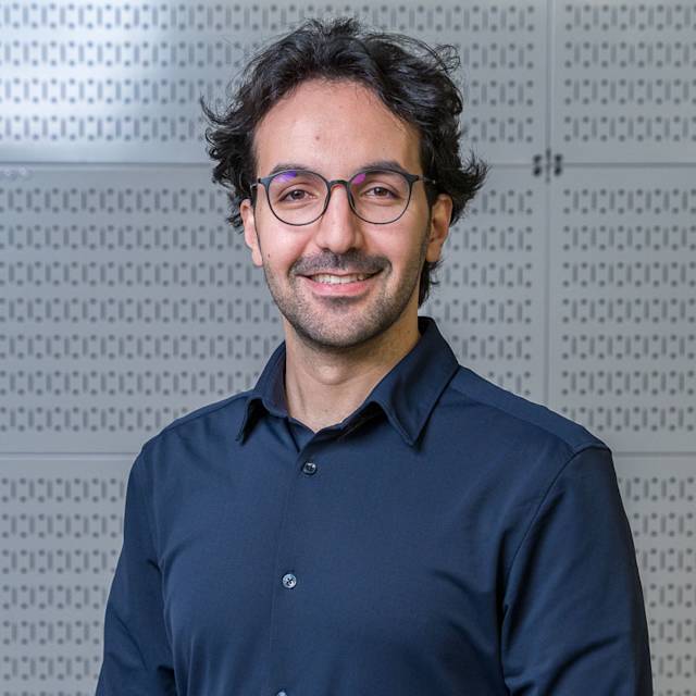 A person with glasses and a beard smiles while standing in front of a perforated metal wall. They are wearing a dark button-up shirt.